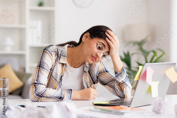 Obraz A tired young Indian girl gets angry from the number of tasks, crumples up the paper and throws it away, tries to calm down by meditating. Stressful work and study, overtasking, headache.