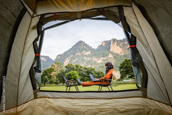 Obraz Young woman sitting near tent with bucket hat hanging on camping chair, using laptop and working remotely while camping in nature during adventure trip in forest. View of hills covered by forest. 
