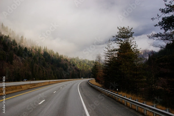 Obraz Asphalt road in the middle of high mountains, covered with fog and clouds on autumn day. American winter landscape of a mountainous area covered with fir forest. Fall season on the highway with cars