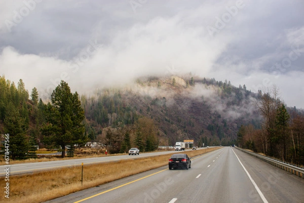 Fototapeta Asphalt road in the middle of high mountains, covered with fog and clouds on autumn day. American winter landscape of a mountainous area covered with fir forest. Fall season on the highway with cars