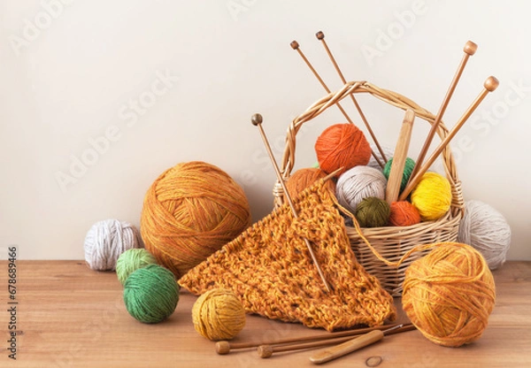 Fototapeta Winter crafts. Process of hand knitting warm bright scarf for cold season. Knitting supplies, set of colorful wool balls in wicker basket, wooden knitting needles and hooks on table. Knitting as hobby