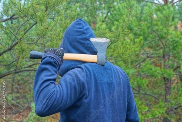 Fototapeta one aggressive man and a bandit in a blue stylish fashionable raglan and a blue hood on his head holds in his hand a small iron new hatchet with a wooden handle behind his back before an attack  

