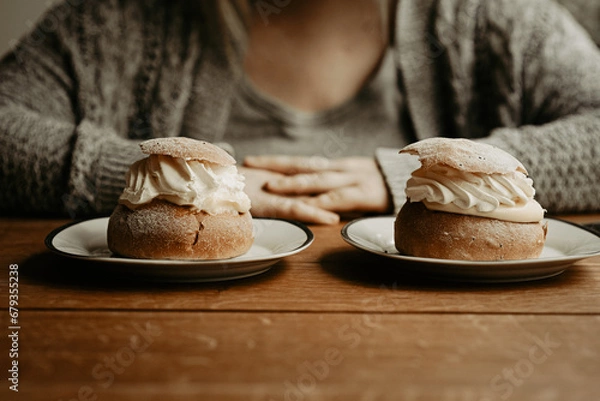 Fototapeta Concept of picking pastry to eat. Focus on traditional Swedish pastry, semla, on the plates at wooden table. In the background, out of focus, a person with hands on table. Photo taken in Sweden.