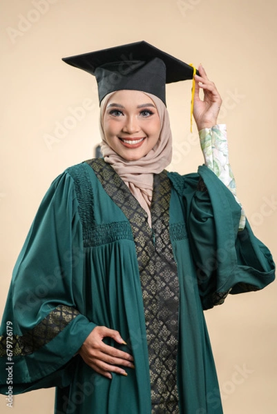 Fototapeta A female muslim lady wearing hijab and convocation attire smiling, isolated on brown background