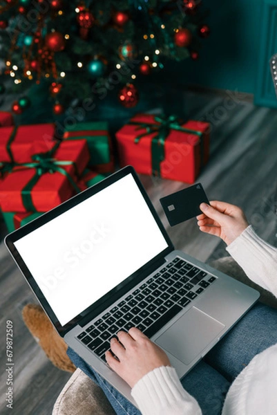Fototapeta Young woman holding a credit card and a gift box against the background of Christmas decor and gifts, close-up. Christmas and New Year shopping on the Internet, payment by credit card