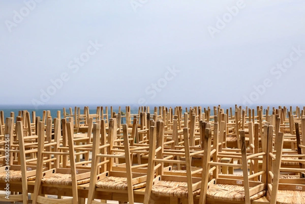 Obraz End of season at the seaside concept background. Large group of wooden chairs of closed seafront outdoor cafe or restaurant placed upside down on top of tables, blurry sea horizon in the distance.