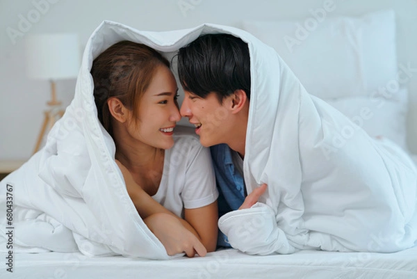 Fototapeta Young Asian couple lovers use noses touch together with love, smiling faces and happiness in bed while using blanket cover heads.