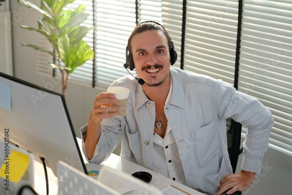 Fototapeta Portrait of Hispanic office worker sitting at desk with headset while holding a cup of hot coffee, smiling face when finish communication. Call center agent support customer to provide information.