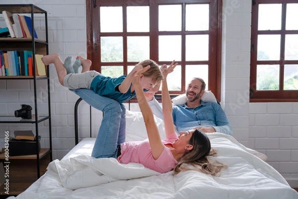 Fototapeta Happy family, mother lying on bed, lifting happy boy in air on straight legs at home. Laughing son feels excited and happy when playing together with daddy and mommy, child flying like plane