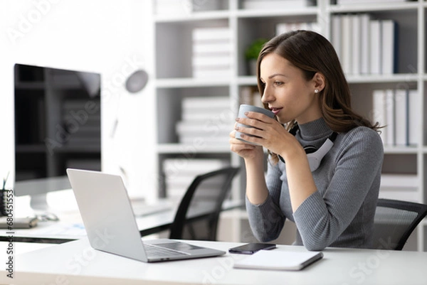 Fototapeta Attractive woman drinking coffee while working on laptop in office at home in morning.