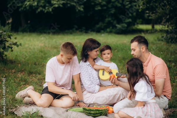 Fototapeta Big family sitting on the picnic blanket in city park during weekend Sunday sunny day. They are smiling, laughing and eating boiled corn and watermelon.