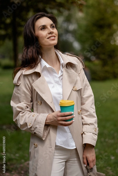 Fototapeta A charming young girl relaxes in a spring park outdoors with a cup of coffee after study or work. A smiling positive lady model within a pensive look is posing in full height for snapshots of