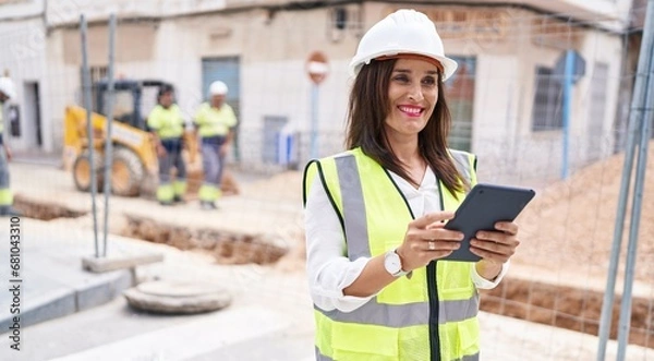 Fototapeta Young beautiful hispanic woman architect using touchpad at street