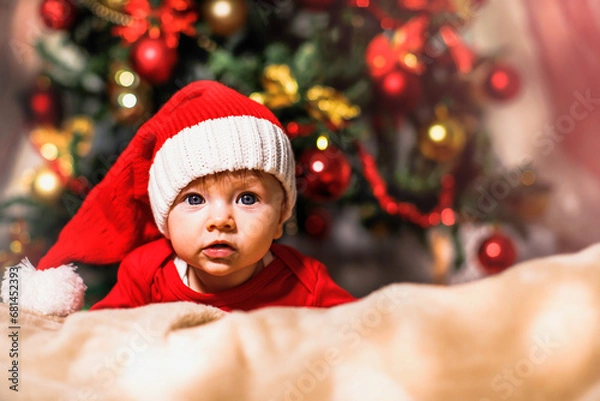 Fototapeta Joyful baby looking at camera in Christmas photo. Newborn baby in red cap of Santa Claus and Christmas tree in background