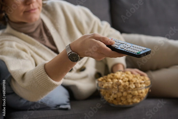 Fototapeta Close-up of young woman lying on sofa and using remote control to watch TV with popcorn