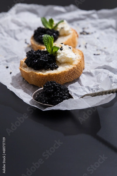 Fototapeta A delicious snack with wine or vodka. Unusual taste combinations, fine dining, wealthy eating concept. Crispy canapes with black caviar, soft cream cheese and sprigs of mint, silver spoon of caviar. 