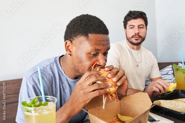 Obraz Friends eating takeout food at table man taking bite of burger