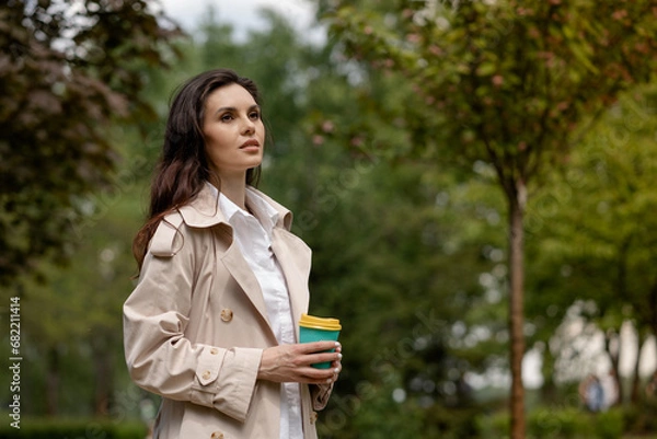 Fototapeta A girl brunette walks in the spring park and holds a cup of coffee in hands. A lady with serious gaze looks into the distance thoughtfully. A young woman enjoys the beauty and harmony of nature alone
