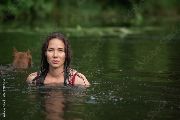 Fototapeta A young beautiful dark-haired girl bathes in a pond with a pit bull terrier dog.