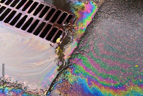 Fototapeta An oil slick against the backdrop of an asphalt road flows into a storm drain through a grate.