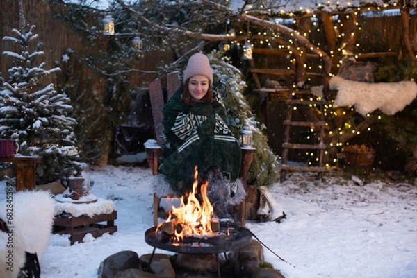 Fototapeta Young attractive woman in winter clothes sitting in a armchair, warming herself by the fire and relaxing outside in winter