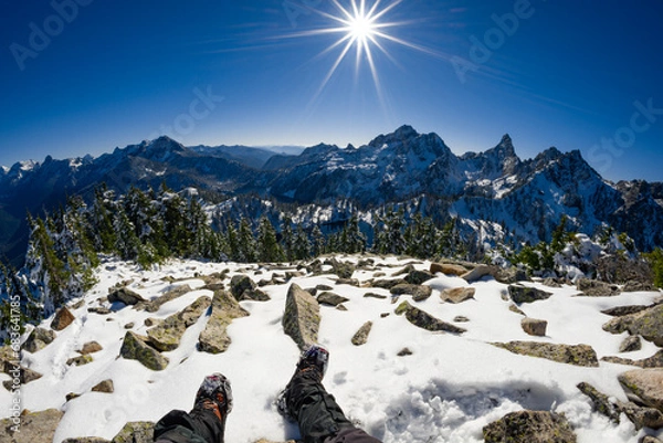 Fototapeta Adventurous athletic male hiker, sitting in a snowy boulder field with his legs outstretched before jagged snowy mountains on a beautiful sunny day.