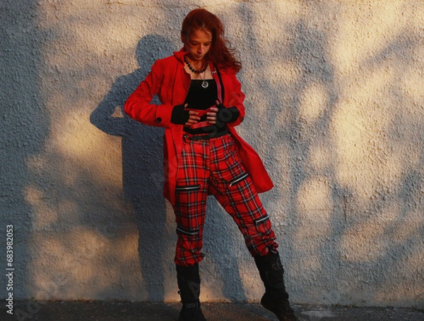 Obraz Selfie photo of a woman near a white gray wall in tight black clothes, a red raincoat, a leather bag. Cinematic punk style, fashion.