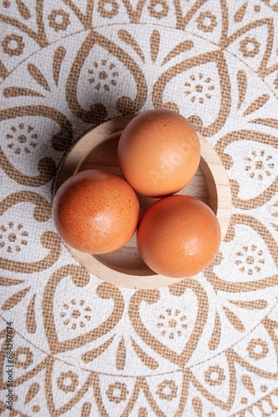 Obraz eggs, aerial photo of three eggs placed on a circular wooden bowl, the table has a tablecloth with mandalas in white and brown tone