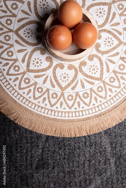 Obraz eggs, aerial photo of three eggs placed on a circular wooden bowl, the table has a tablecloth with mandalas in white and brown tone