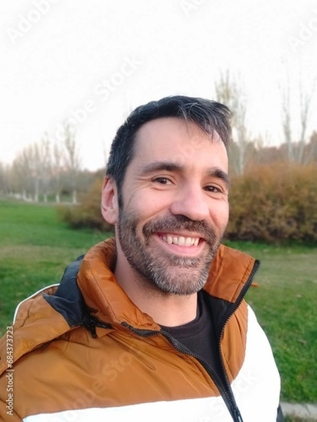 Fototapeta selfie of black haired caucasian man smiling with short beard and orange and white jacket in a park