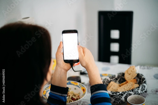 Obraz Woman from behind with dark hair looking at the phone screen in a family environment, with food in front of the table in the background.