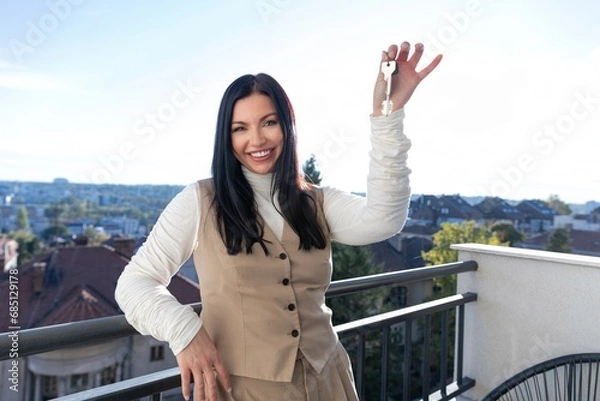 Fototapeta Girl holding keys in hand to new apartment on background of houses and smiling, concept of affordable housing and mortgage