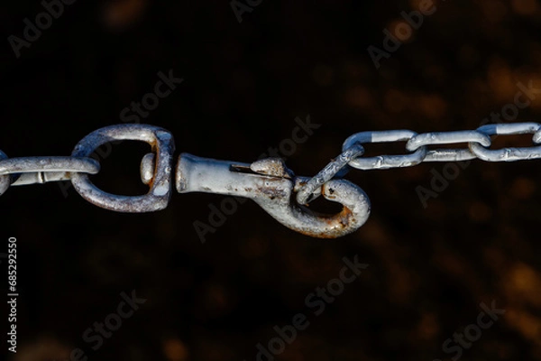 Fototapeta "A close-up of the metal hook of a dog leash attached to an old chain with aged and whitened links against a dark background."