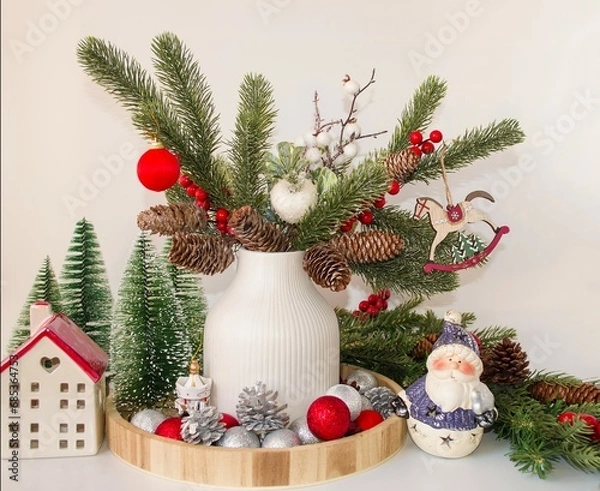 Obraz Beautiful holiday card.  Merry Christmas and New Year greeting concept.  On a white background, a vase with decorated fir branches, a ceramic house, cute Santa Claus.  Foreground.