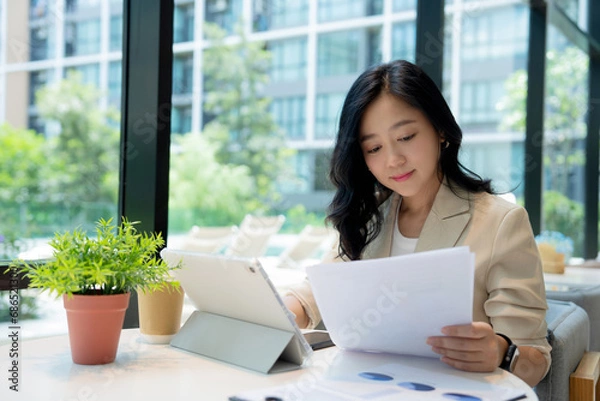 Fototapeta Beautiful Asian businesswoman analyzing charts using calculator and laptop with documents and holding a pen.