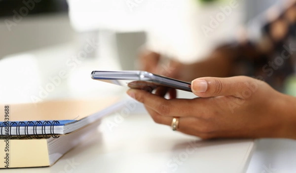 Fototapeta Student writes in notebook and holds smartphone. Woman hands taking notes using online organizer app on mobile phone concept
