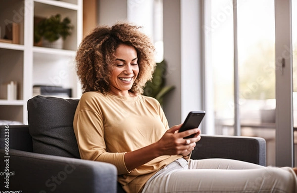 Fototapeta Happy casual beautiful woman holding mobile phone sitting on a sofa at home. Smiling woman checking social media using a  smartphone. 