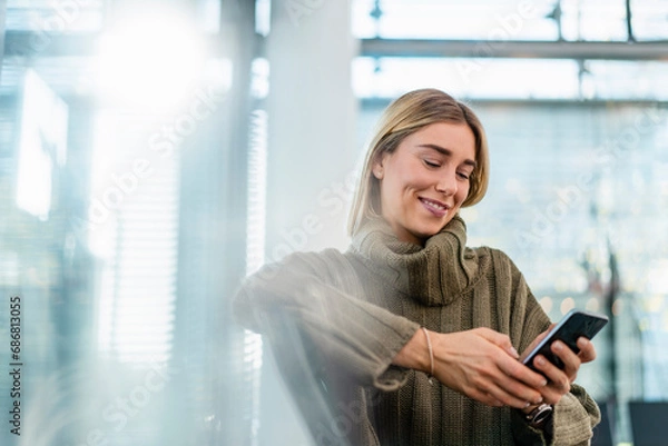 Obraz Smiling young woman sitting in waiting area using cell phone