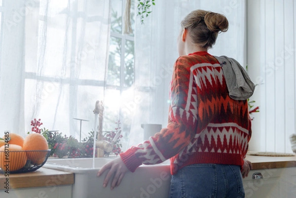 Obraz young woman in red sweater stays in kitchen leaning on sink feeling upset. thoughtful and frustrated girl at home during homework looks through window at Christmas holidays