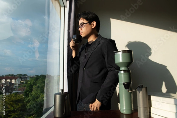 Obraz asian man wear black suit dark shade glasses stand in fornt of desk books make confidence mood and coffee with city window view and shadow on wall