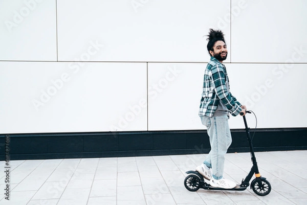 Fototapeta Trendy smiling bearded man in casual clothes riding electric scooter in urban background. Handsome model posing in the street near wall. Hipster guy with curly hairstyle. Cheerful and happy