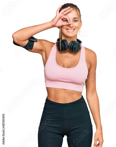 Fototapeta Young blonde woman wearing gym clothes and using headphones doing ok gesture with hand smiling, eye looking through fingers with happy face.