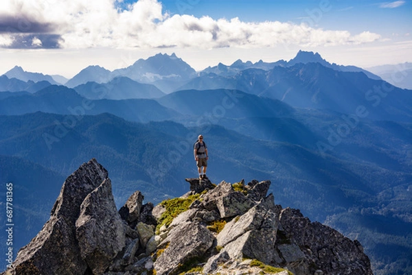 Fototapeta Adventurous athletic male hiker standing on top of a rugged mountain in the Pacific Northwest with jagged mountains in the background.
