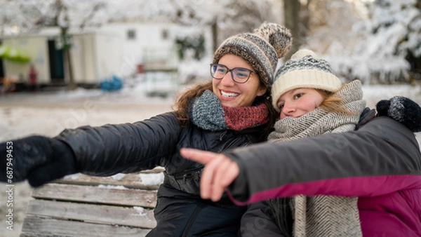 Fototapeta Joyful Winter Moments: Two Young Women in Wool Hats and Scarfs Pointing and Laughing on Snowy Bench