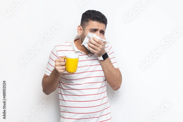 Fototapeta Portrait of sick unhealthy bearded man wearing striped t-shirt standing suffering runny nose, drinking hot tea, needs treatment. Indoor studio shot isolated on gray background.