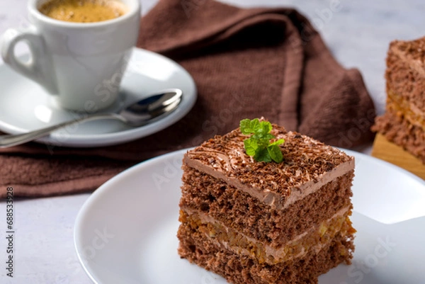 Fototapeta Chocolate cake cut into square pieces with nut cream. Dessert on a white plate with coffee