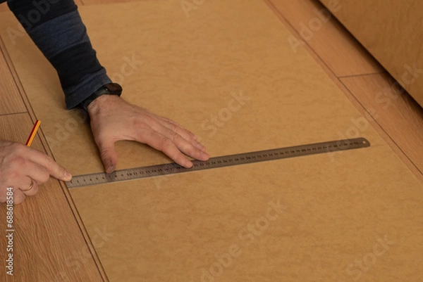 Fototapeta A man measures a wooden board with a ruler and makes marks with a pencil. Assembling a wooden product. Human hands and plywood.