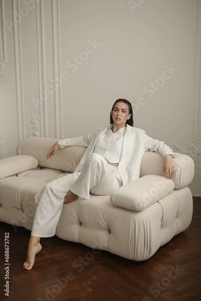 Obraz A confident brunette woman dressed in a white pinstripe women's business suit is posing on a small white couch in a photo studio. The studio interior features white walls and a wooden parquet floor.