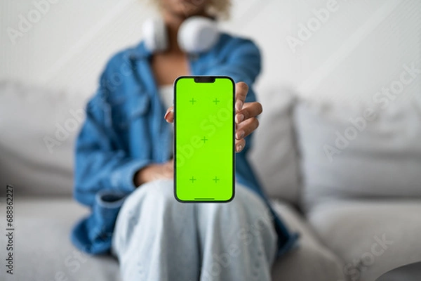 Obraz Young Woman holding Green Mock-up Screen Smartphone. She's Sitting On a Couch in His Cozy Living Room. Mobile applications for food delivery,online services etc