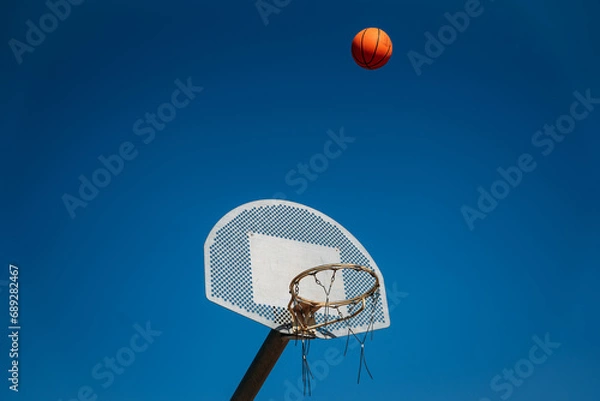 Fototapeta Basketball basket seen from below and one side while an orange ball is falling from above. Contrasted with a blue sky on a sunny, clear day.